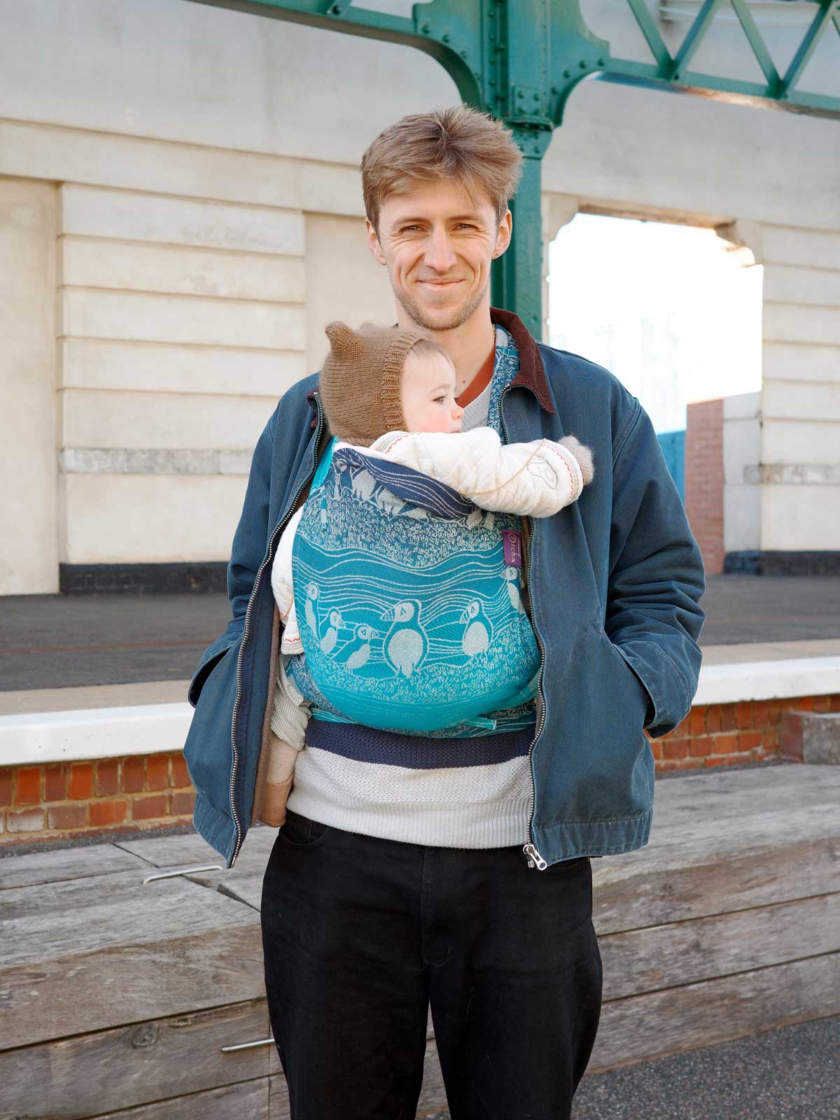Man carrying a baby in a blue sling outdoors.