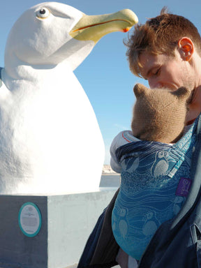 Person carrying a baby in a carrier next to a large white bird sculpture with a blue sky background