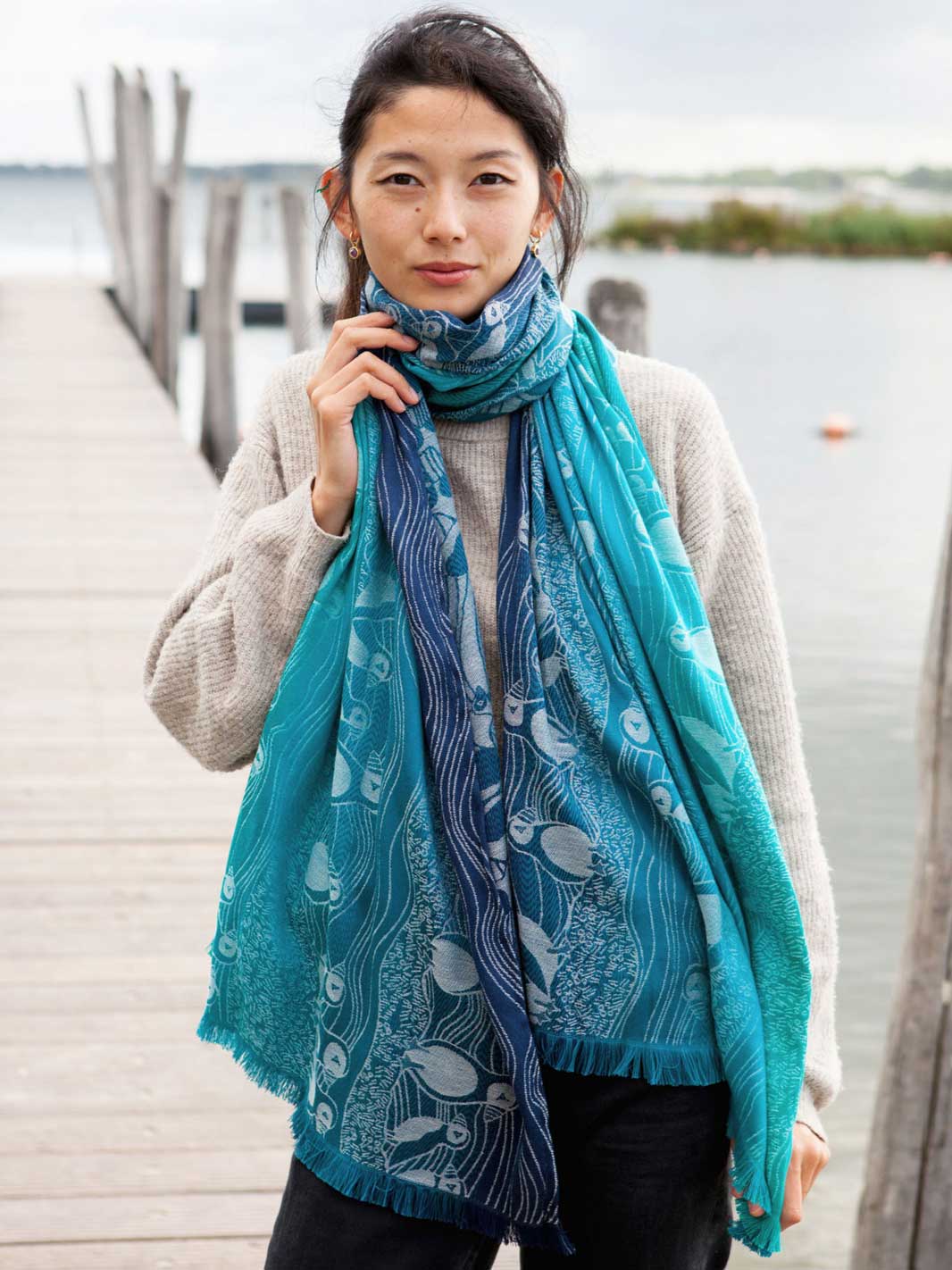 A woman standing on a pier wearing a teal and blue scarf with puffin pattern