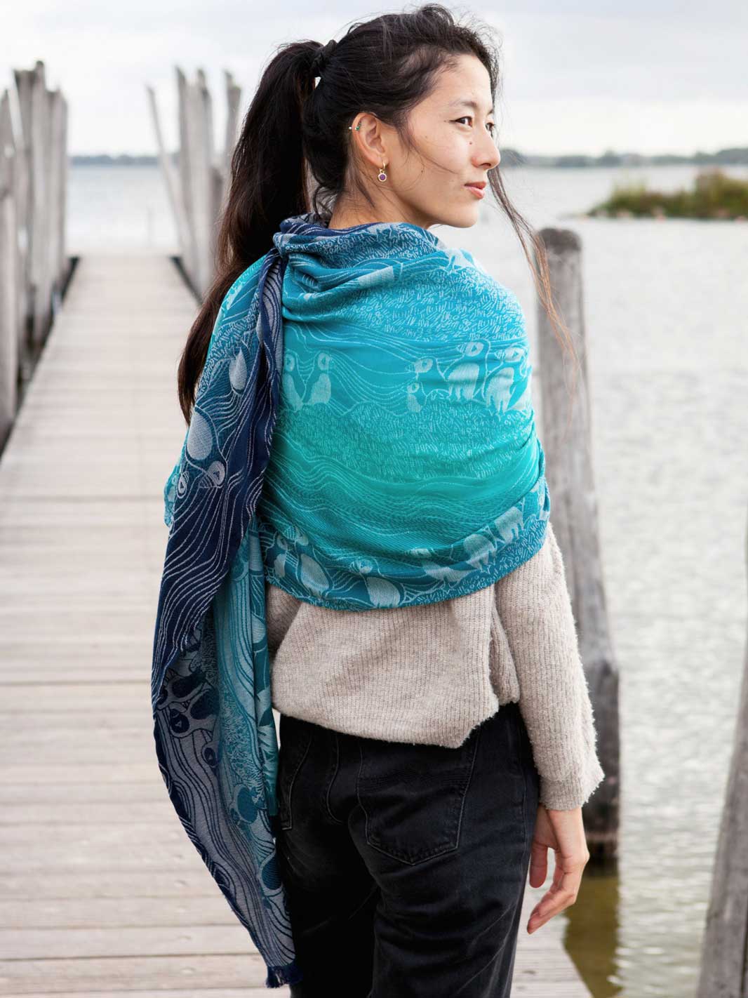 Rear view of a woman standing on a pier wearing a teal and blue scarf with puffin pattern like a shawl