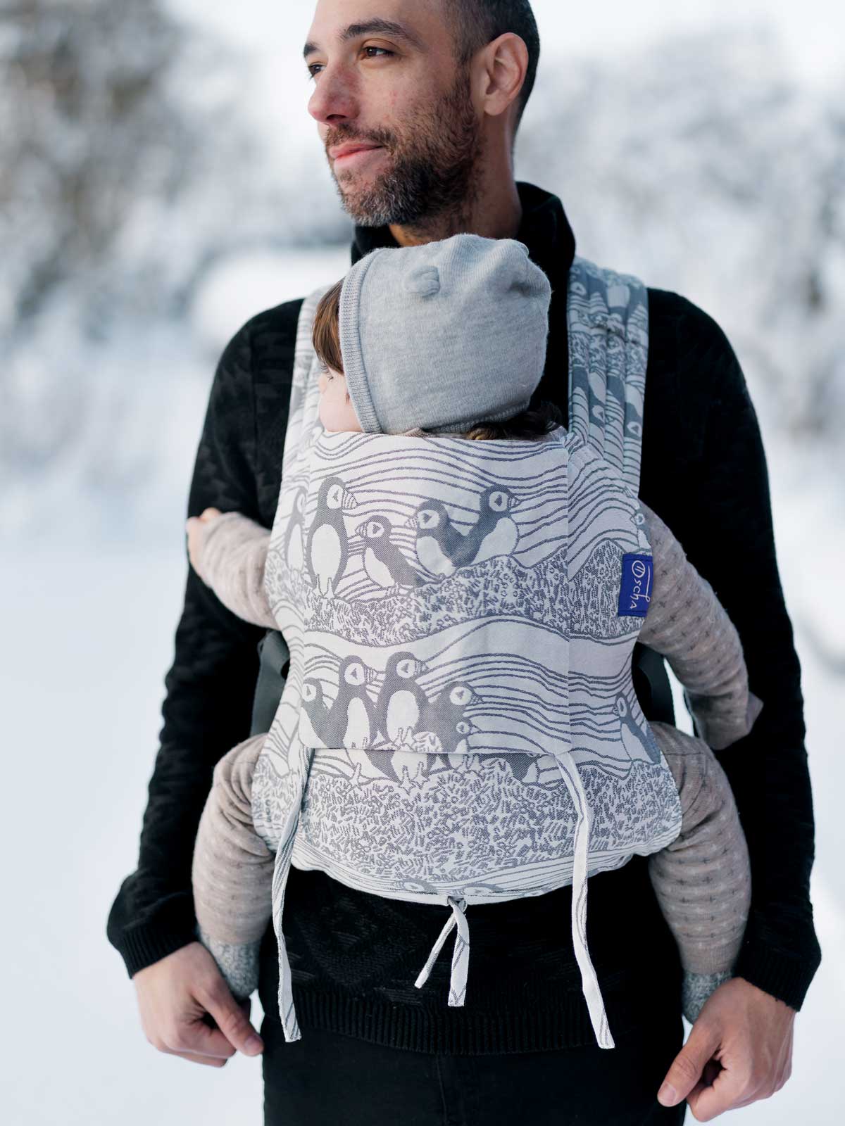 Man carrying a child in a baby carrier with a puffin design against a snowy background