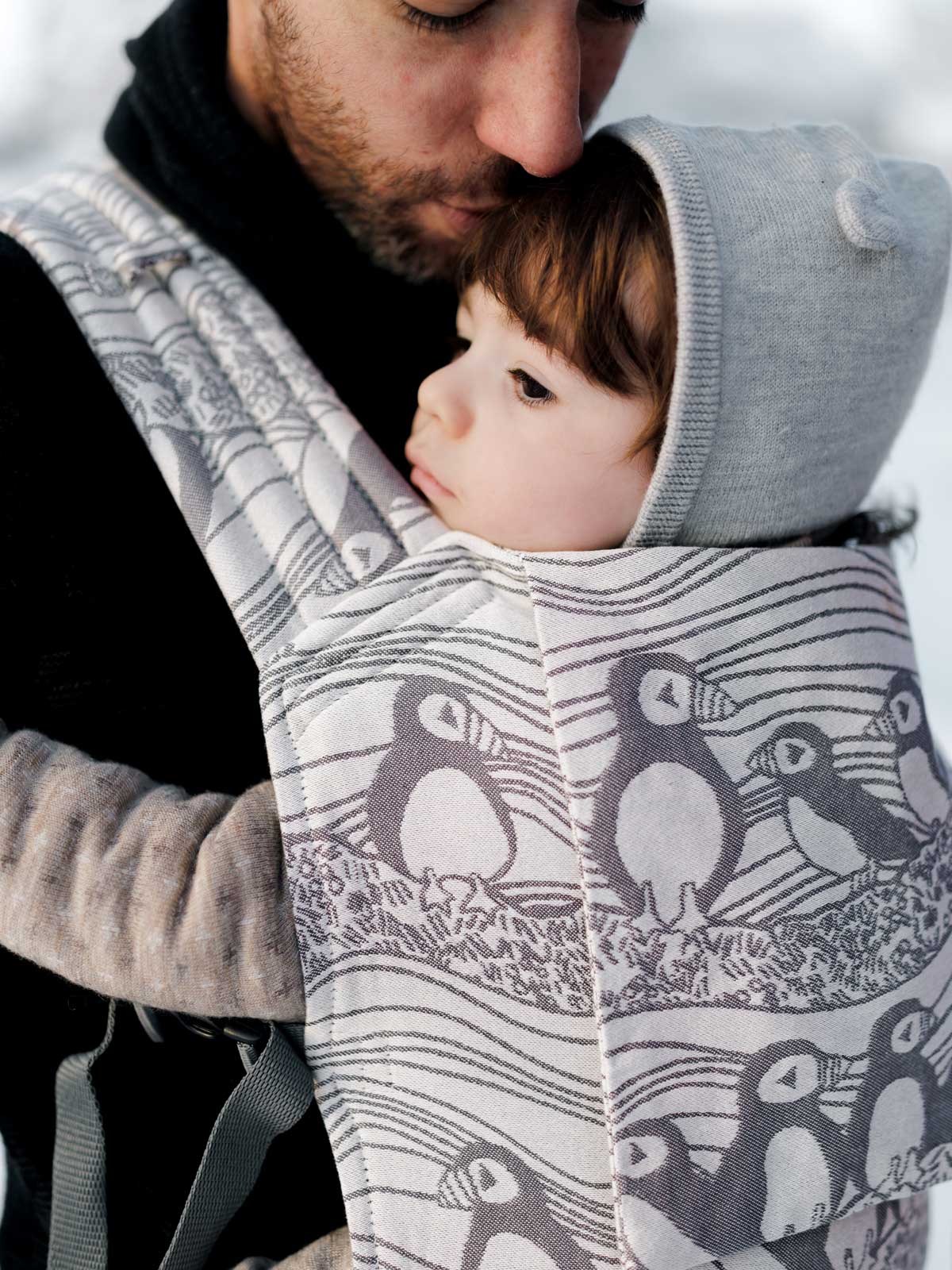 Person wearing a baby in a carrier with puffin design, set against a snowy background