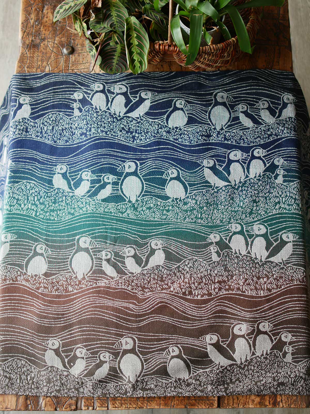 Blue, green and brown fabric with puffin design on a wooden table with plants in background.