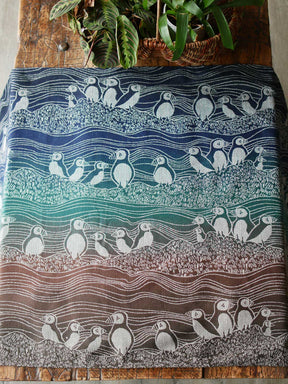 Blue, green and brown fabric with puffin design on a wooden table with plants in background.