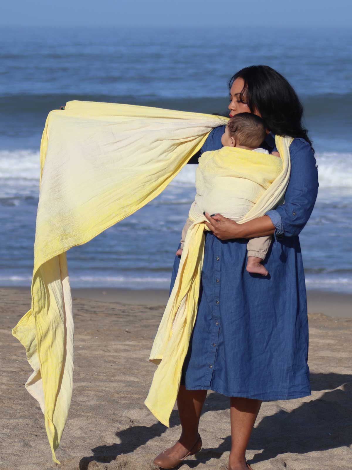 Woman putting a baby in a yellow woven wrap on a beach