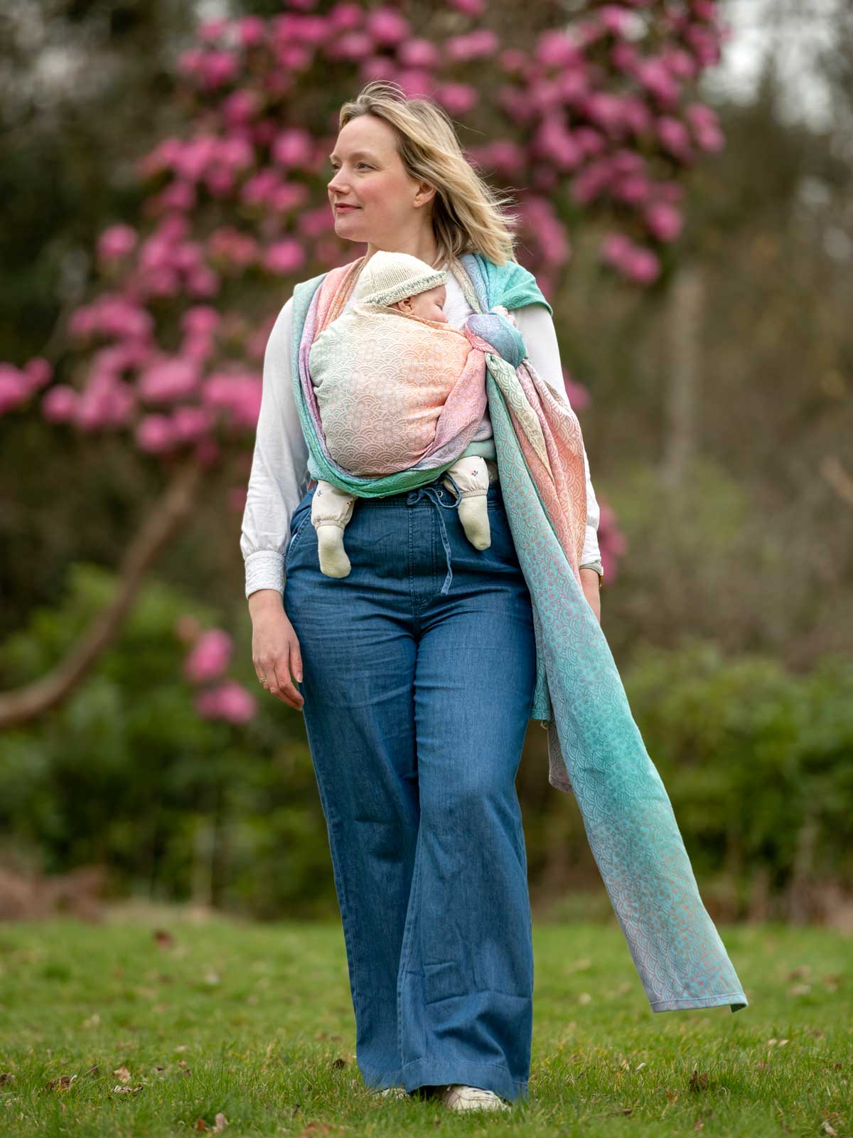 Woman walking outdoors with a baby in a sling, surrounded by pink flowers and greenery.