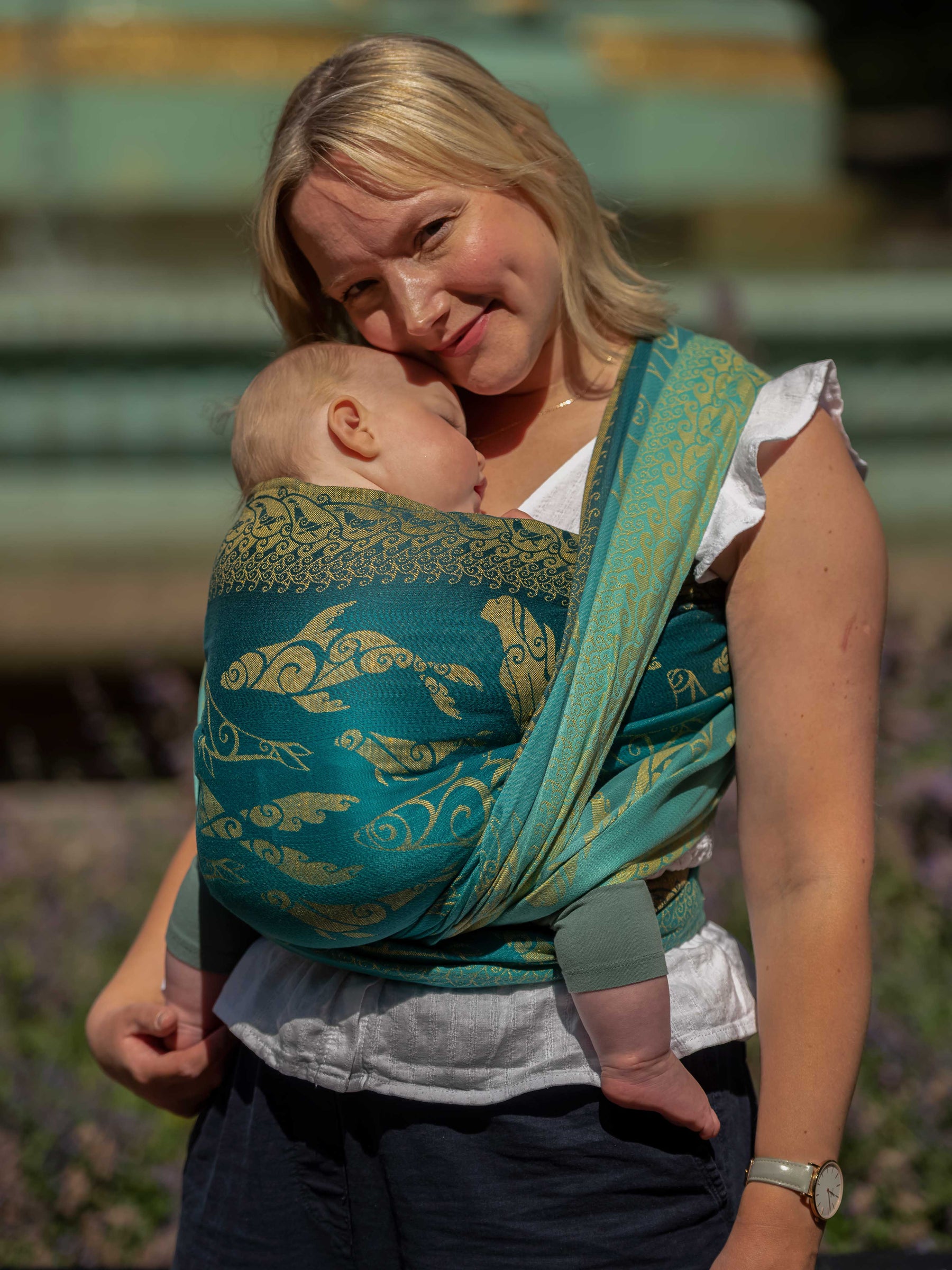 Side view of a woman carrying a baby in a teal woven wrap with gold Selkies