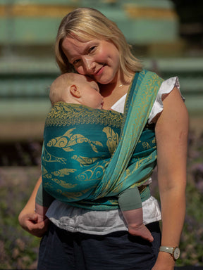 Side view of a woman carrying a baby in a teal woven wrap with gold Selkies