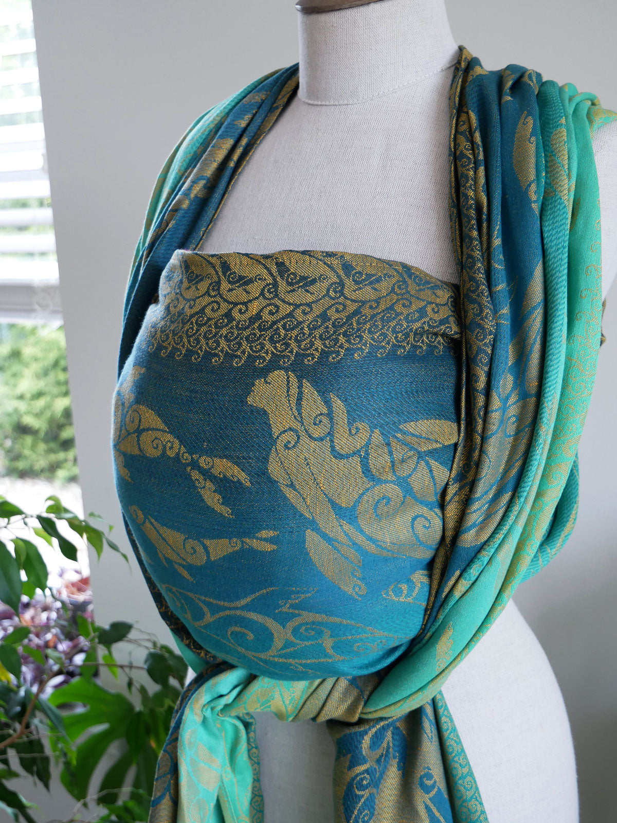 Mannequin wearing a blue and green patterned Selkies Sounds Baby Wrap with a blurred indoor background