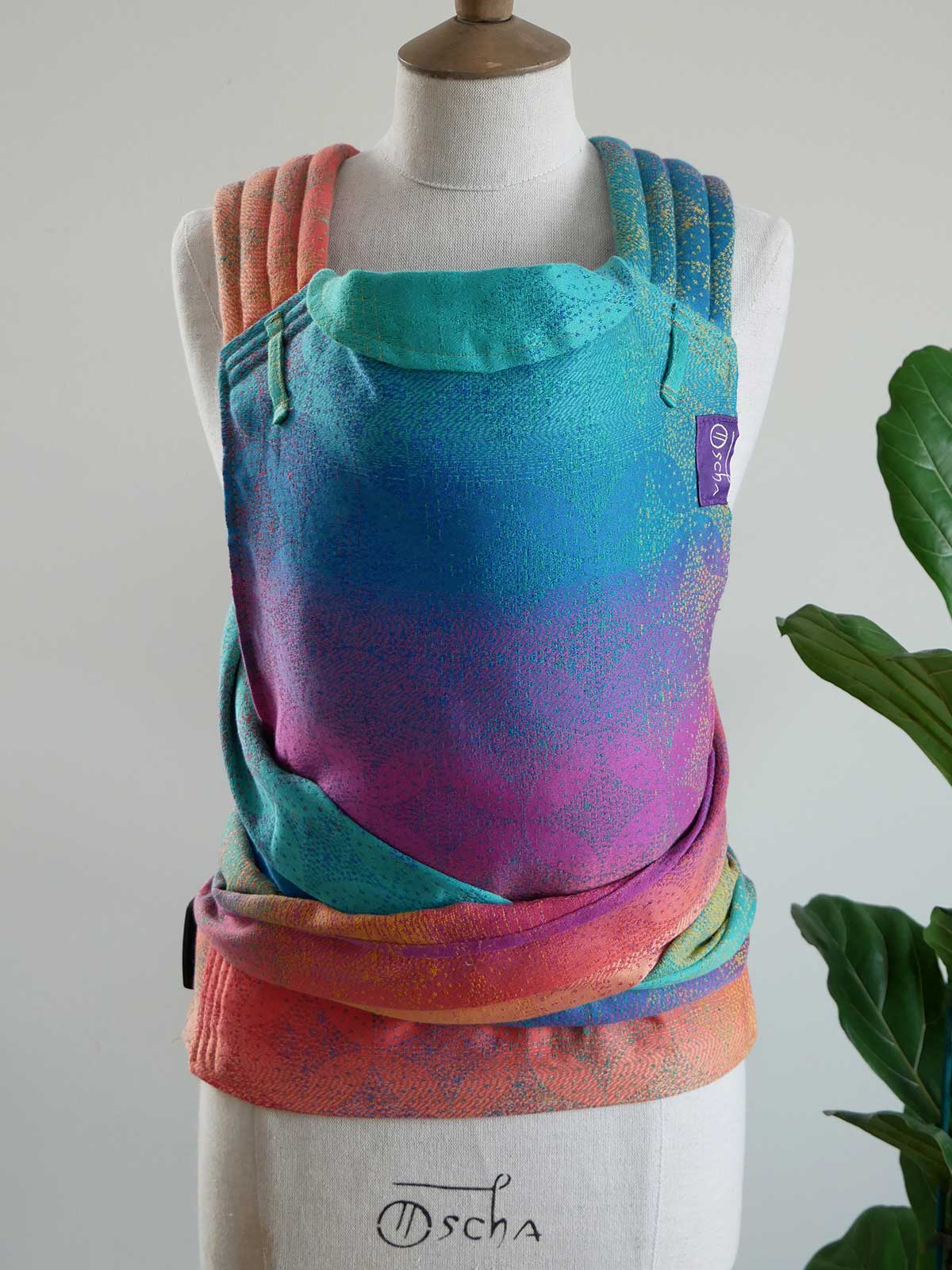 Colorful baby carrier on a mannequin with 'Oscha' branding against a neutral background.