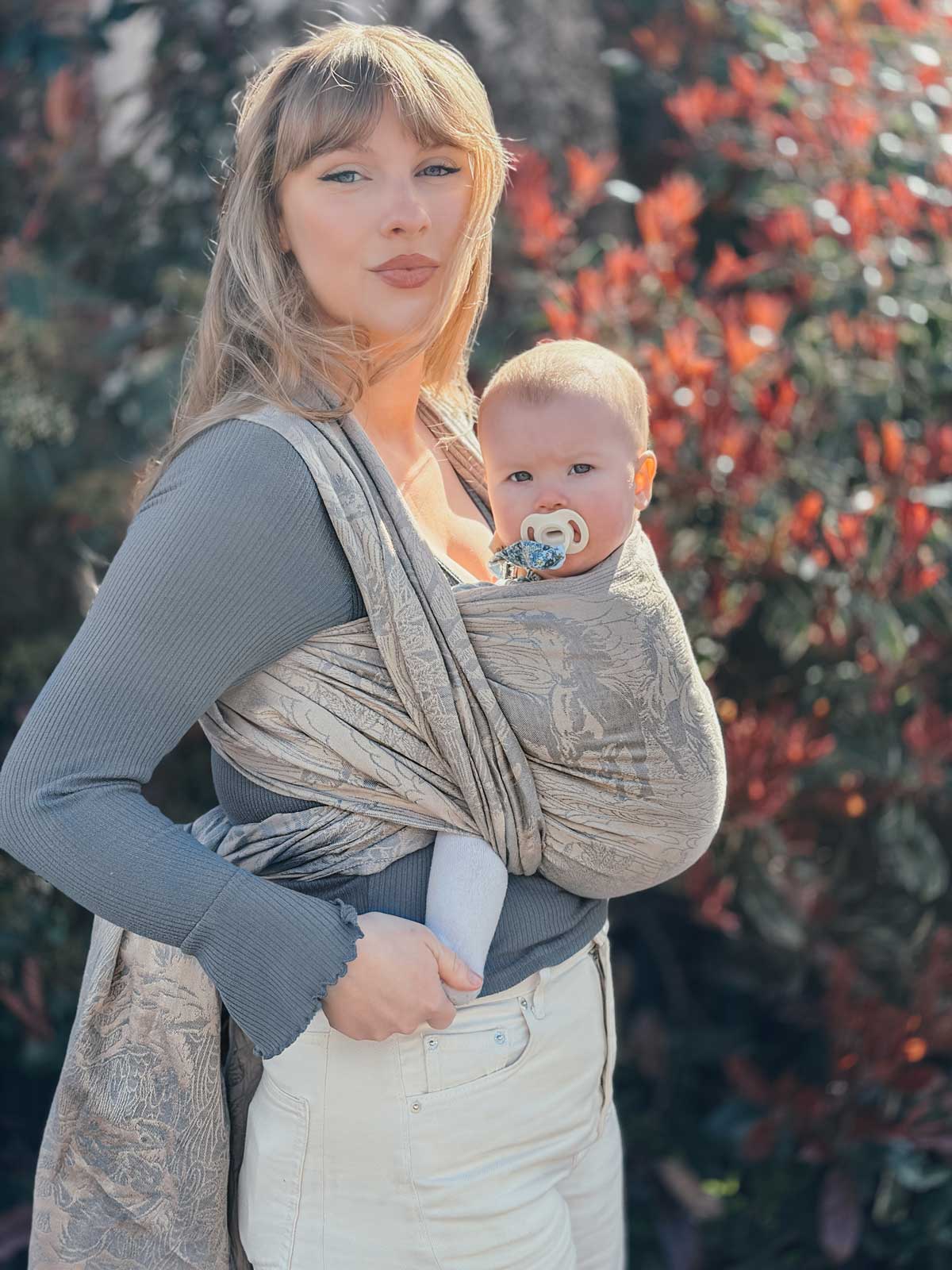 Woman holding a baby in a sling outdoors with blurred foliage in the background
