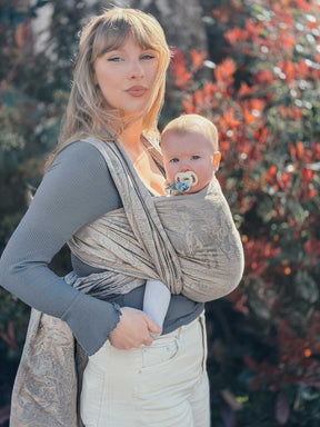 Woman holding a baby in a sling outdoors with blurred foliage in the background