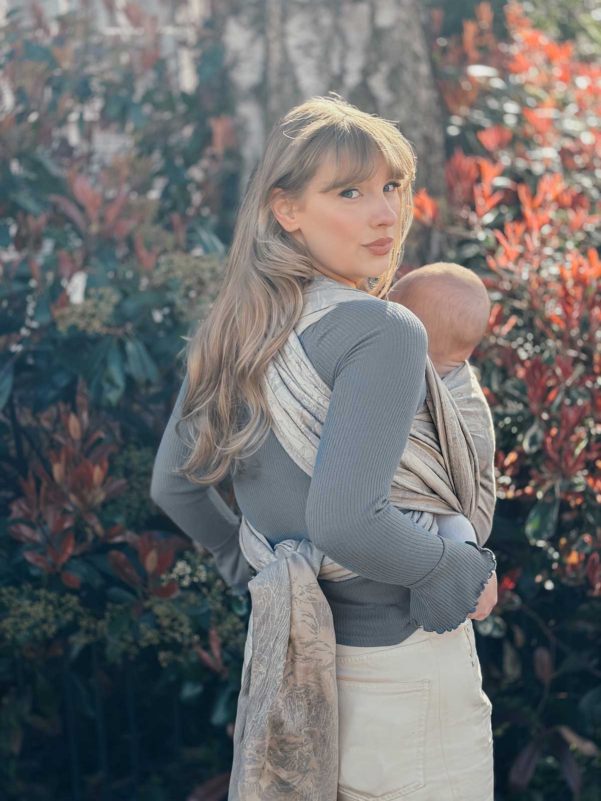 Woman holding a baby in a sling against a natural background with trees and plants.