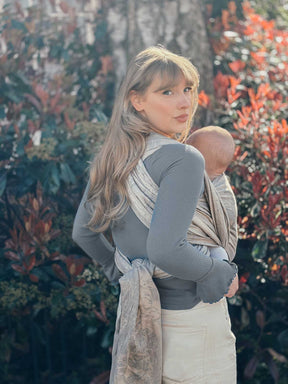 Woman holding a baby in a sling against a natural background with trees and plants.