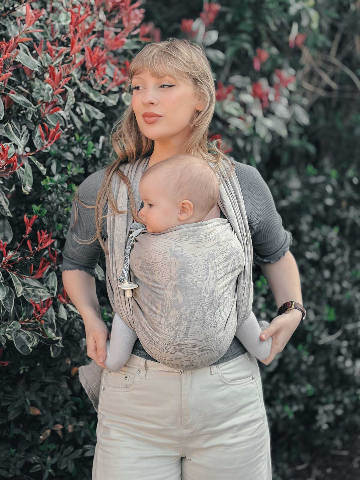 Woman carrying a baby in a sling outdoors with greenery in the background