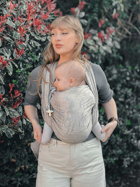 Woman carrying a baby in a sling outdoors with greenery in the background