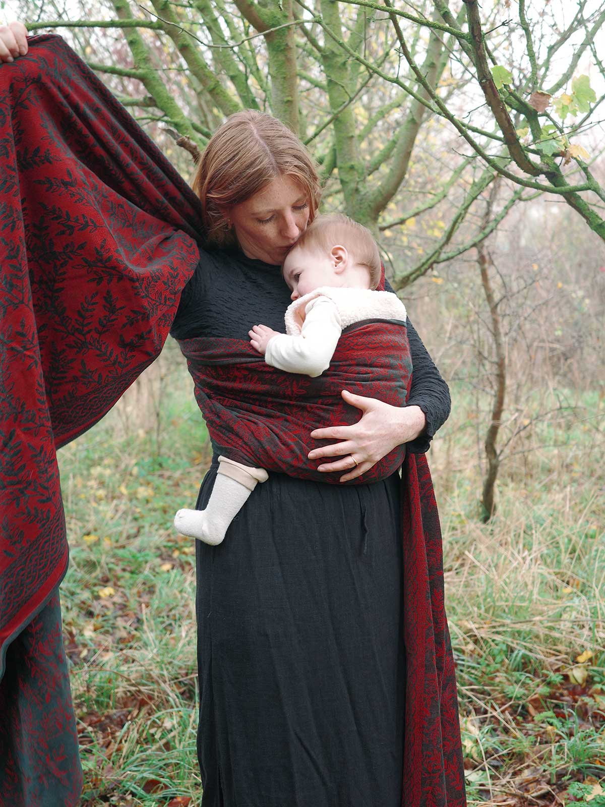 Woman holding a baby in a red wrap outdoors with trees and grass in the background