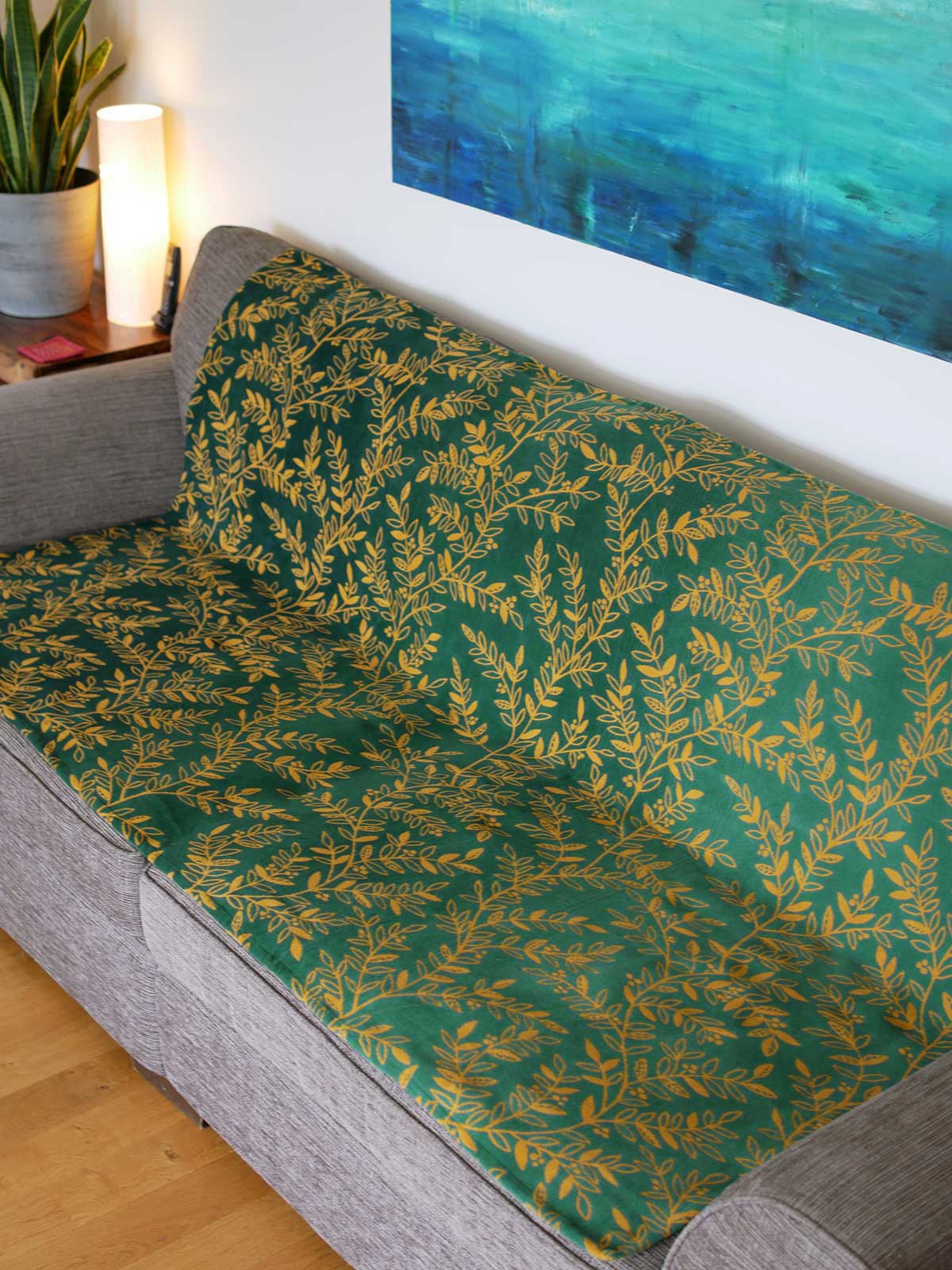 Green throw with golden willow leaves design. The throw sits on a grey sofa.