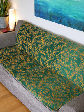 Green throw with golden willow leaves design. The throw sits on a grey sofa.