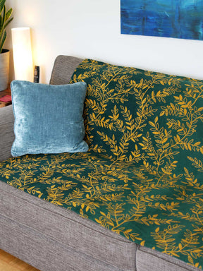 Green throw with golden willow leaves design. The throw sits on a grey sofa.