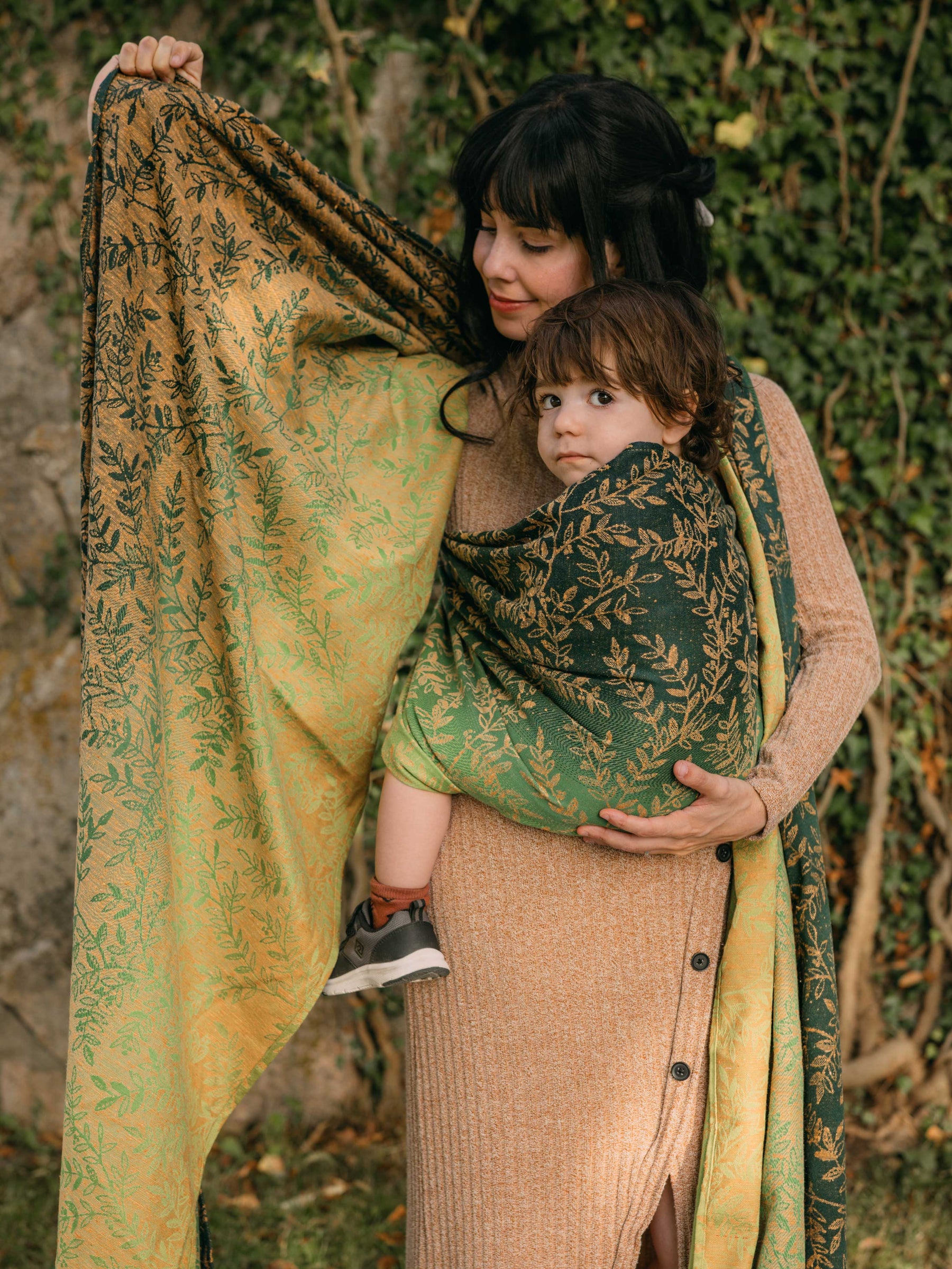 A woman wrapping a child in a green woven wrap with gold leaf pattern