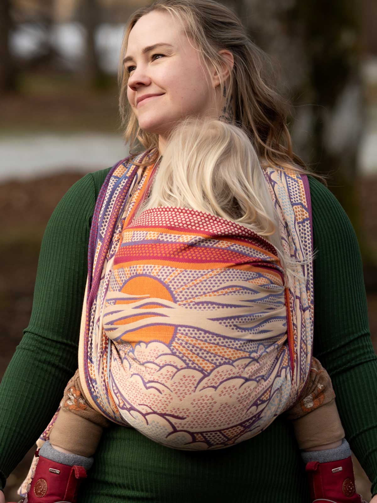 Woman wearing a colorful baby woven wrap with a scenic design outdoors.