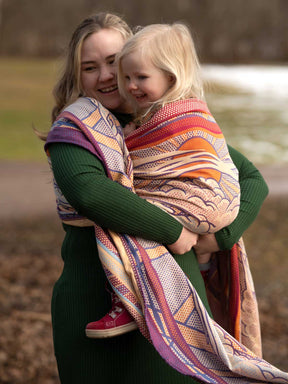 Woman holding a child wrapped in a colorful sling outdoors.